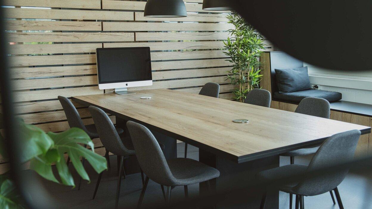 Stilvolle Büroecke mit langem Holztisch, grauen Stühlen und iMac, umgeben von einer Holzlamellenwand und Pflanzen.
