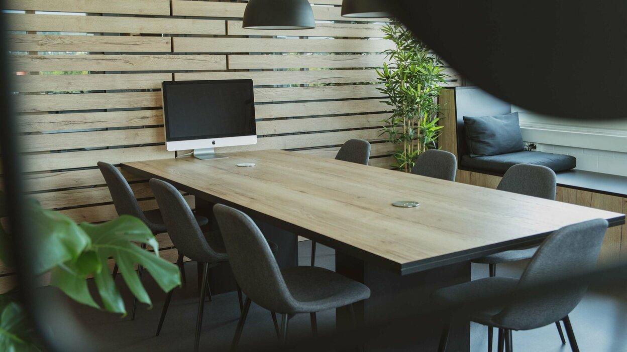 Modernes Interior Design mit Holzelementen Stilvolle Büroecke mit langem Holztisch, grauen Stühlen und iMac, umgeben von einer Holzlamellenwand und Pflanzen.