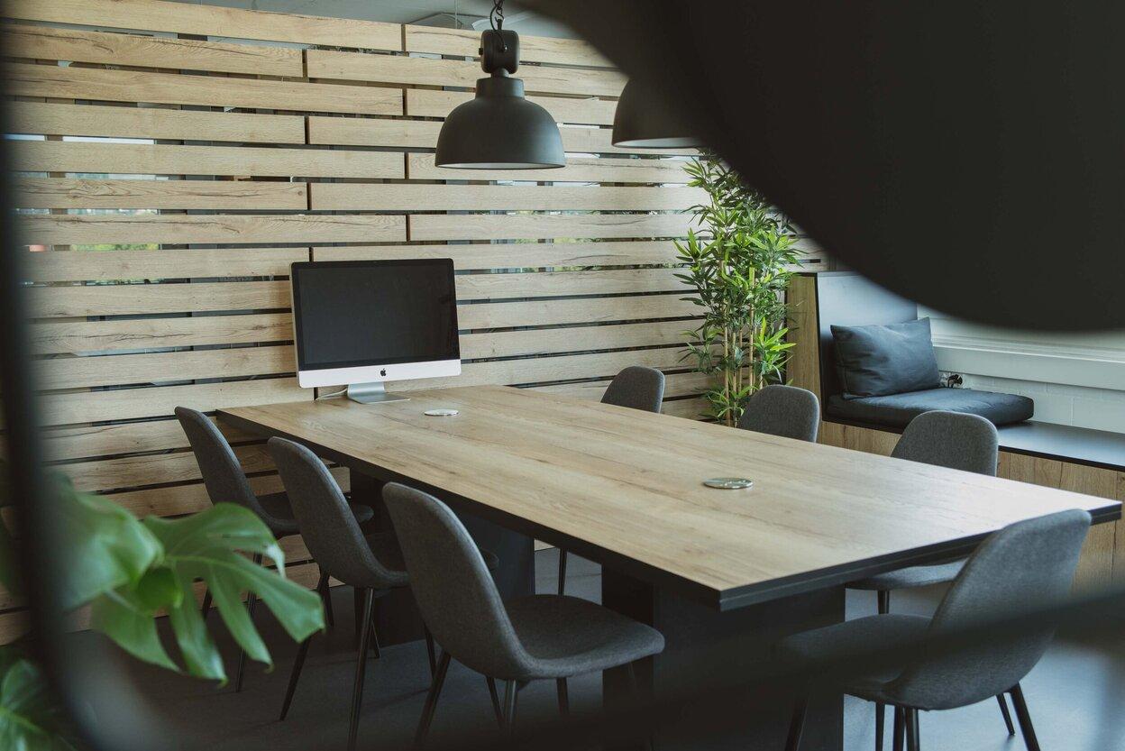 Stilvolle Büroecke mit langem Holztisch, grauen Stühlen und iMac, umgeben von einer Holzlamellenwand und Pflanzen.