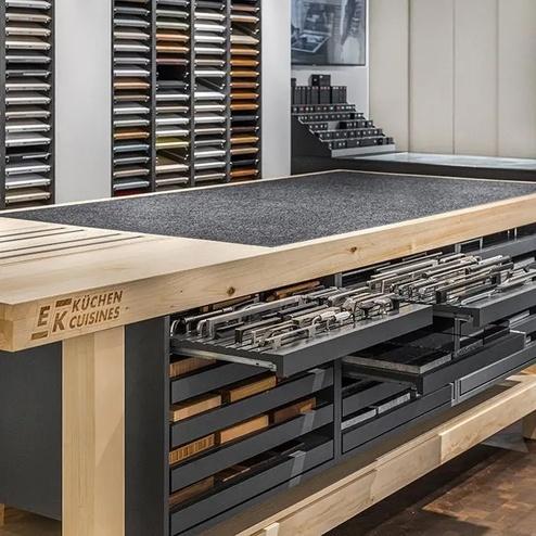 Ausstellungstisch für Küchenplanung mit Material- und Griffmustern Holzarbeitstisch mit Schubladen für Griffe und Materialien, umgeben von Wandregalen mit Küchenfrontmustern in einer Küchenplanungsausstellung.