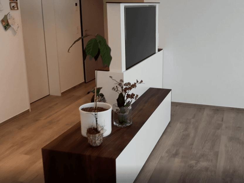 Motorisiertes Fernsehmöbel in Weiss- und Holzdesign mit dekorativen Pflanzen und hochgefahrenem Bildschirm in einem modernen Raum.