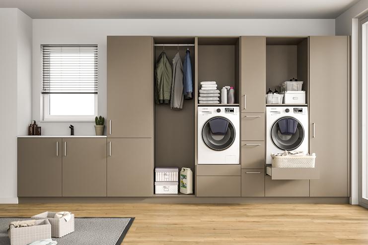 Ein heller Raum mit einem grossen, massgeschneiderten Schranksystem in Taupe, integrierten Waschmaschinen, Ablageflächen und einem Fenster mit Jalousien.